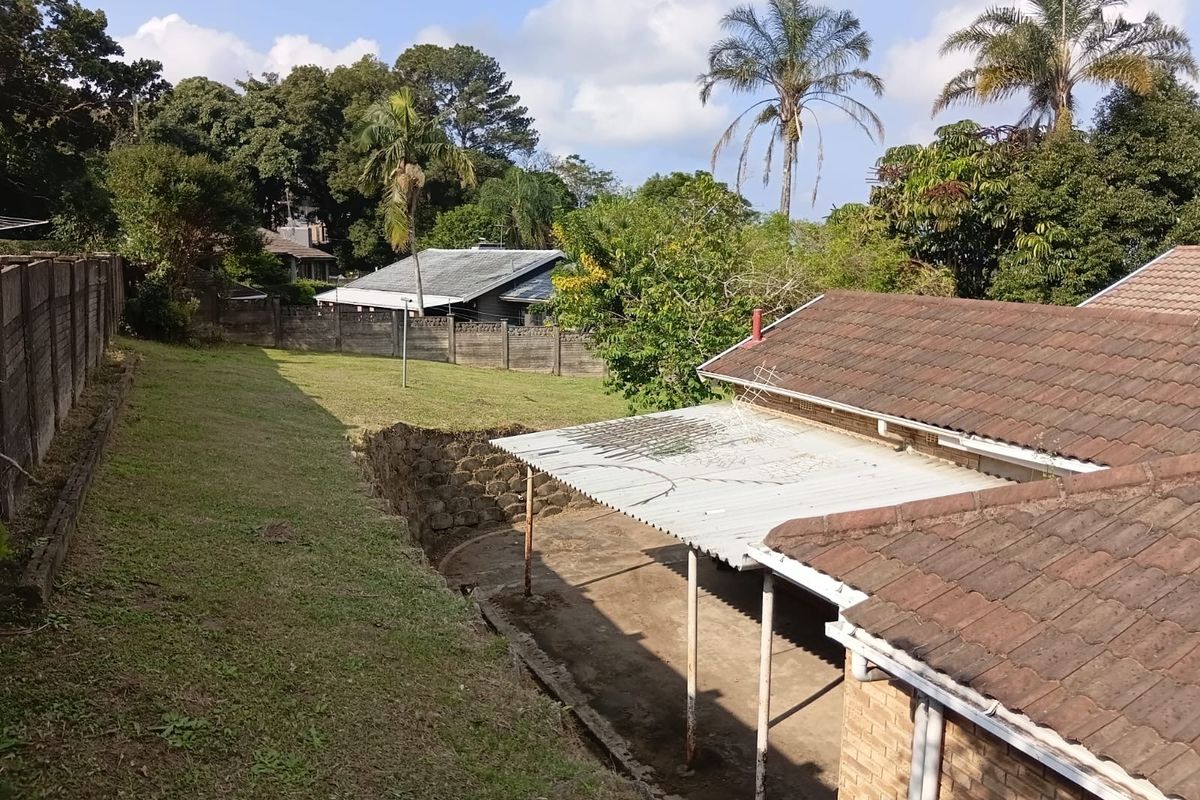 Rear view of the property featuring a tiled roof, a covered patio area with corrugated metal roofing, and a large garden with stone retaining walls.