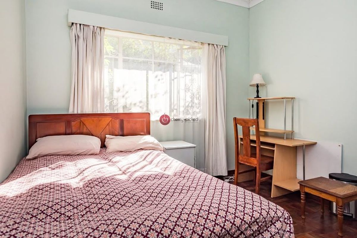 A well-lit bedroom featuring classic parquet flooring, neutral wall tones, and a large window with security bars.