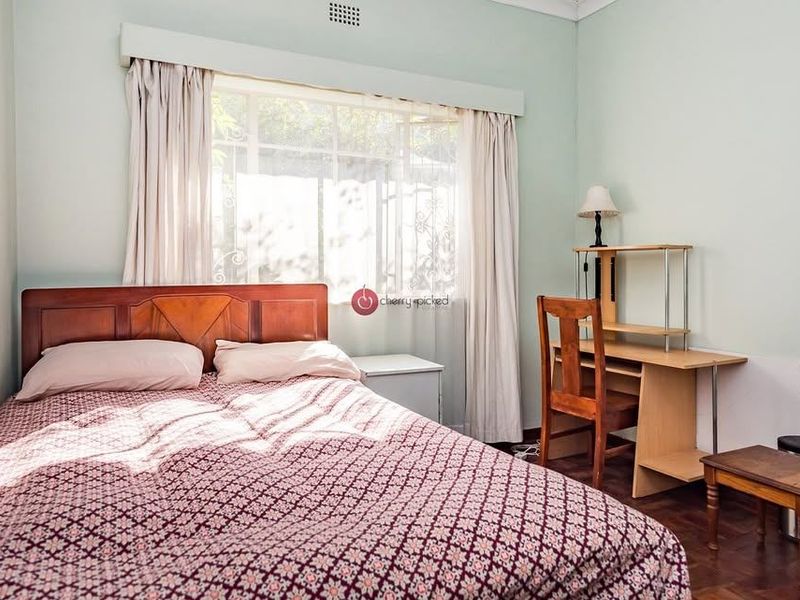 A well-lit bedroom featuring classic parquet flooring, neutral wall tones, and a large window with security bars.