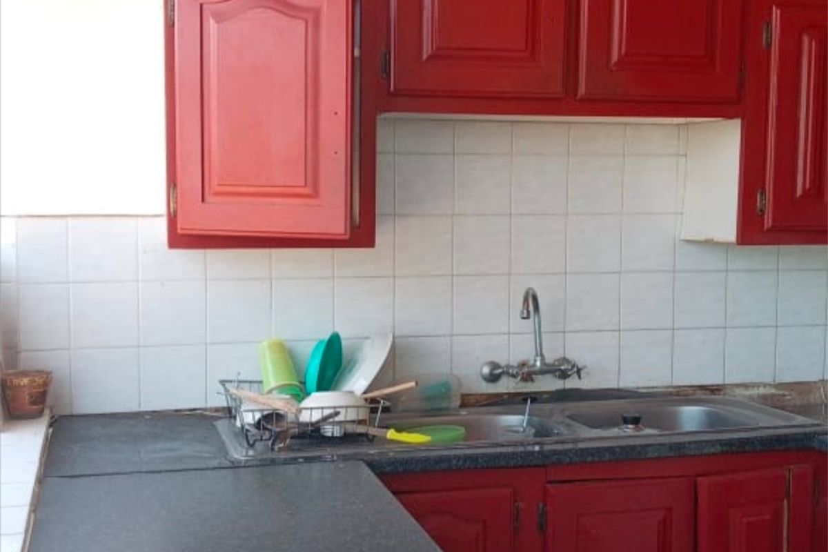 A dated kitchen featuring red cabinetry, a dark laminate countertop, a stainless steel sink, and a built-in electric hob with a tiled backsplash.