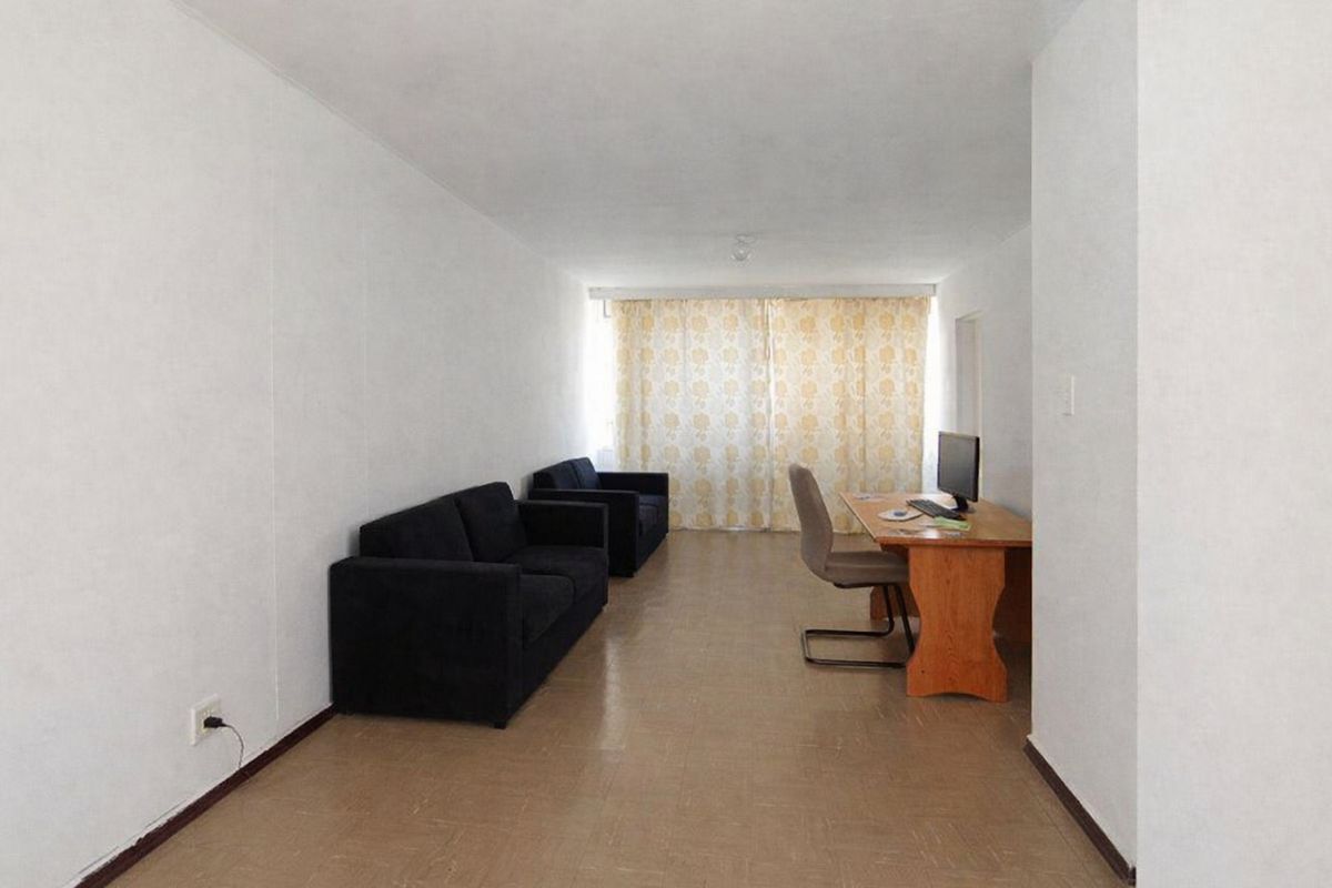 A simple, rectangular living area featuring neutral walls and vinyl flooring, illuminated by natural light through large window curtains.