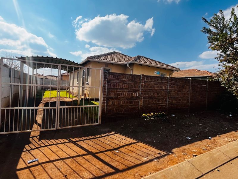 Front view of a residential property featuring a brick boundary wall, metal driveway gate, and a carport structure.