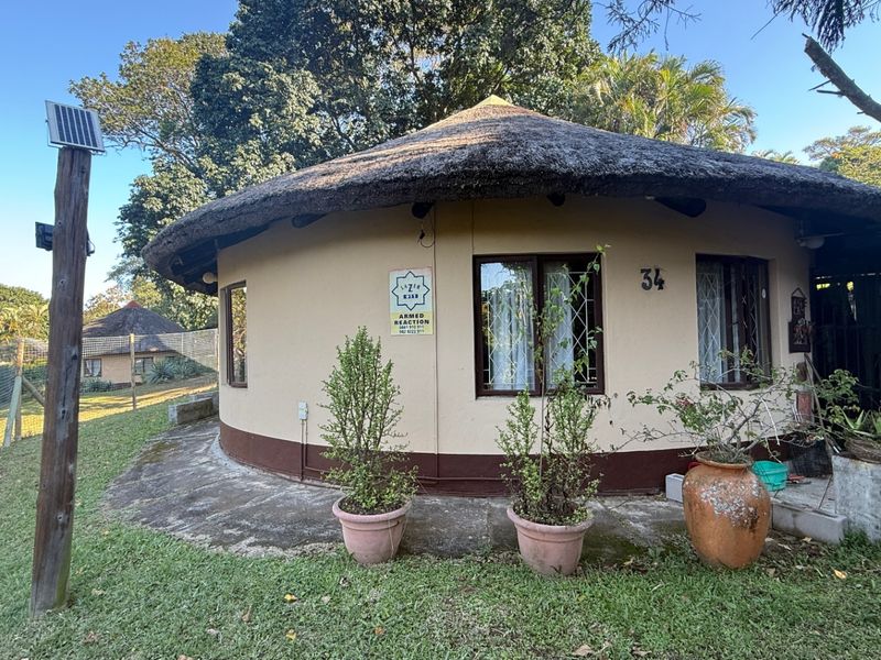 A circular townhouse unit featuring a traditional thatched roof, security-barred windows, and a surrounding concrete apron set within a garden environment.
