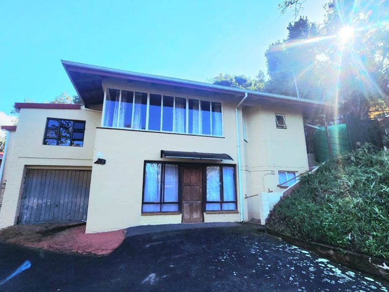 Front elevation of a two-story townhouse featuring a garage, timber entrance door, and large windows, set against a sloped, landscaped embankment.