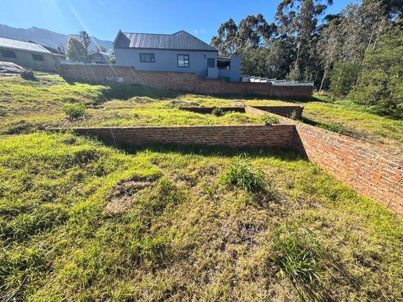 A vacant plot featuring exposed brick foundation footings and retaining walls, set against a backdrop of established residential properties and natural landscape.
