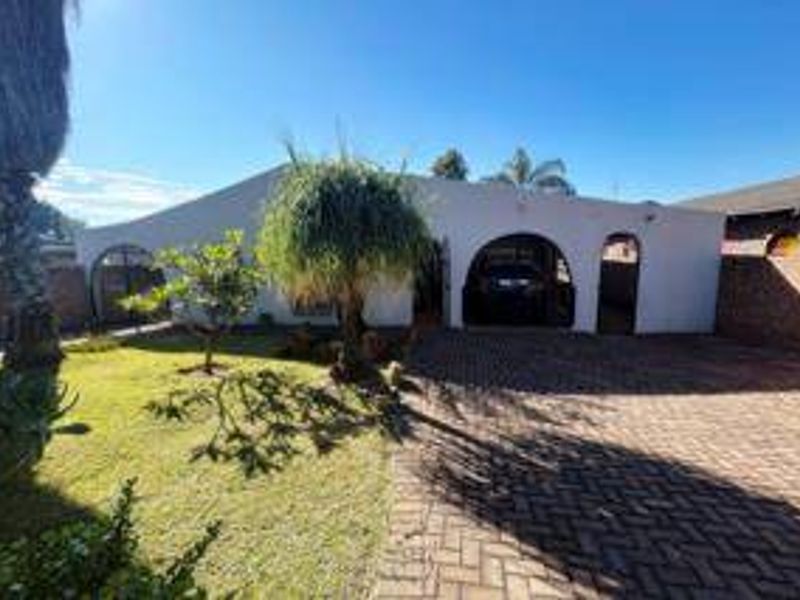 Front elevation of a single-story house featuring a paved driveway, arched carport entrance, and established garden area.
