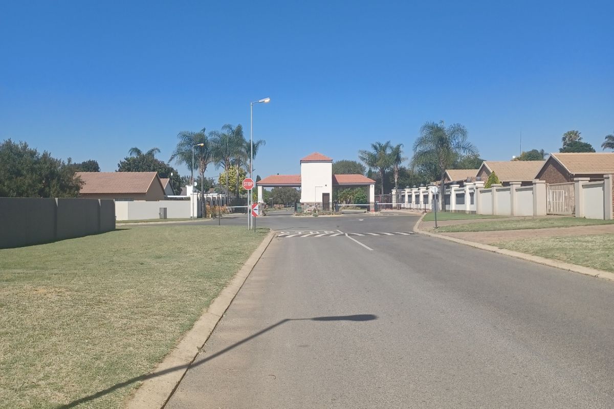 A view of the residential estate entrance featuring a security gatehouse, perimeter walls, and paved access road under a clear blue sky.