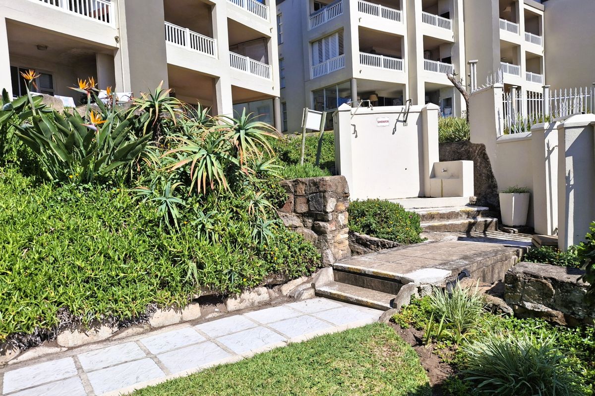 Exterior view of an apartment complex featuring a landscaped garden, paved walkway, and a communal outdoor shower area.