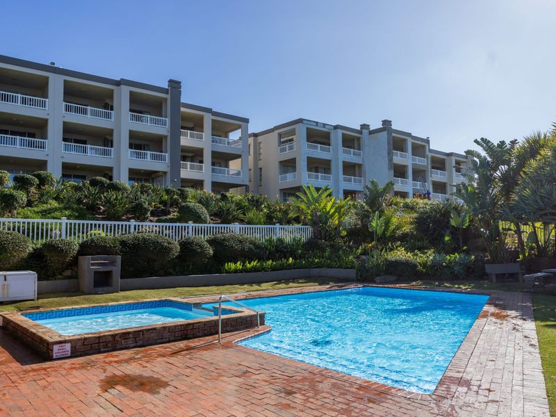 Communal swimming pool and spa area featuring brick paving, built-in braai facilities, and landscaped gardens set against a multi-story apartment complex.