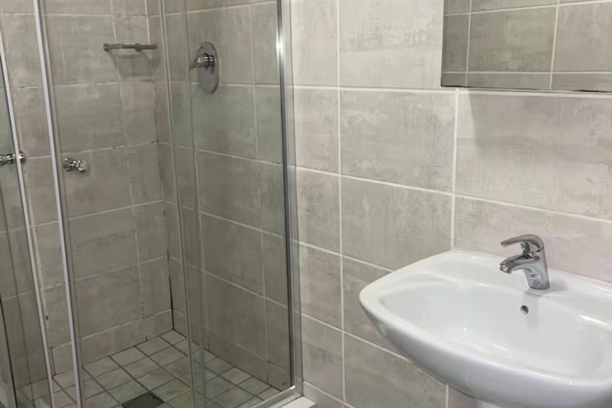 A functional bathroom featuring a glass-enclosed shower, pedestal washbasin, and neutral wall tiling.