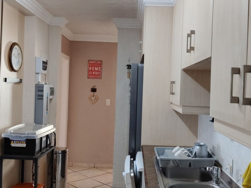 A functional kitchen area featuring light wood-tone cabinetry, laminate countertops, and a stainless steel sink, leading into a tiled hallway.