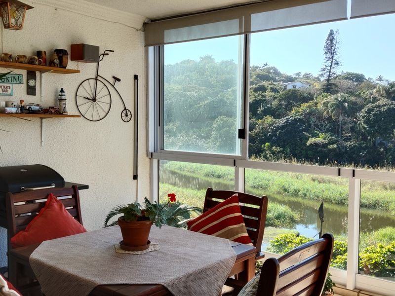 A bright, enclosed balcony featuring large sliding glass windows with roller blinds, offering scenic views of the surrounding greenery and water.