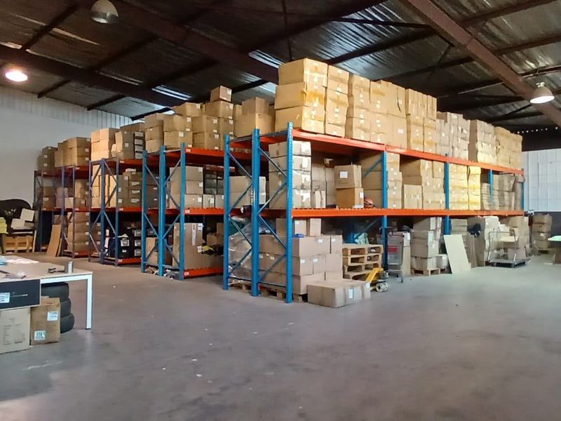 Spacious industrial warehouse featuring high ceilings with exposed steel trusses, concrete flooring, and extensive built-in steel racking systems for storage.