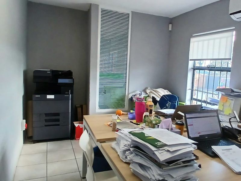 A functional office space featuring tiled flooring, a wall-mounted air conditioning unit, and a window equipped with security bars and a roller blind.