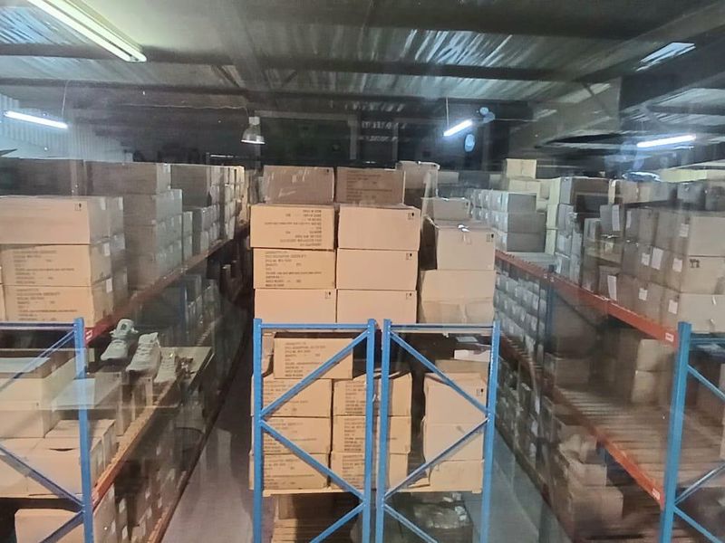 Industrial warehouse storage area featuring extensive metal racking systems, exposed steel roof trusses, and overhead fluorescent lighting.