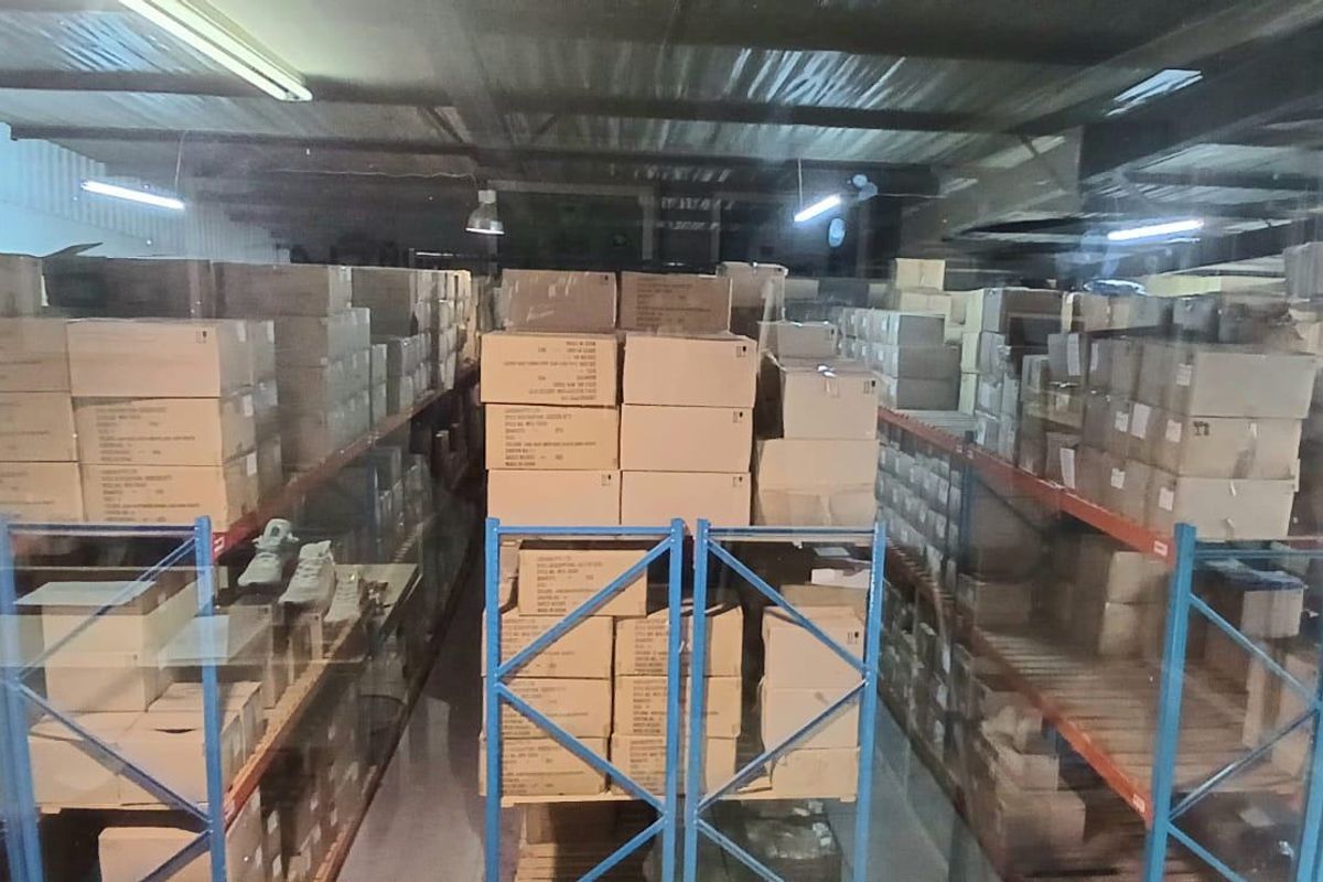Industrial warehouse storage area featuring extensive metal racking systems, exposed steel roof trusses, and overhead fluorescent lighting.