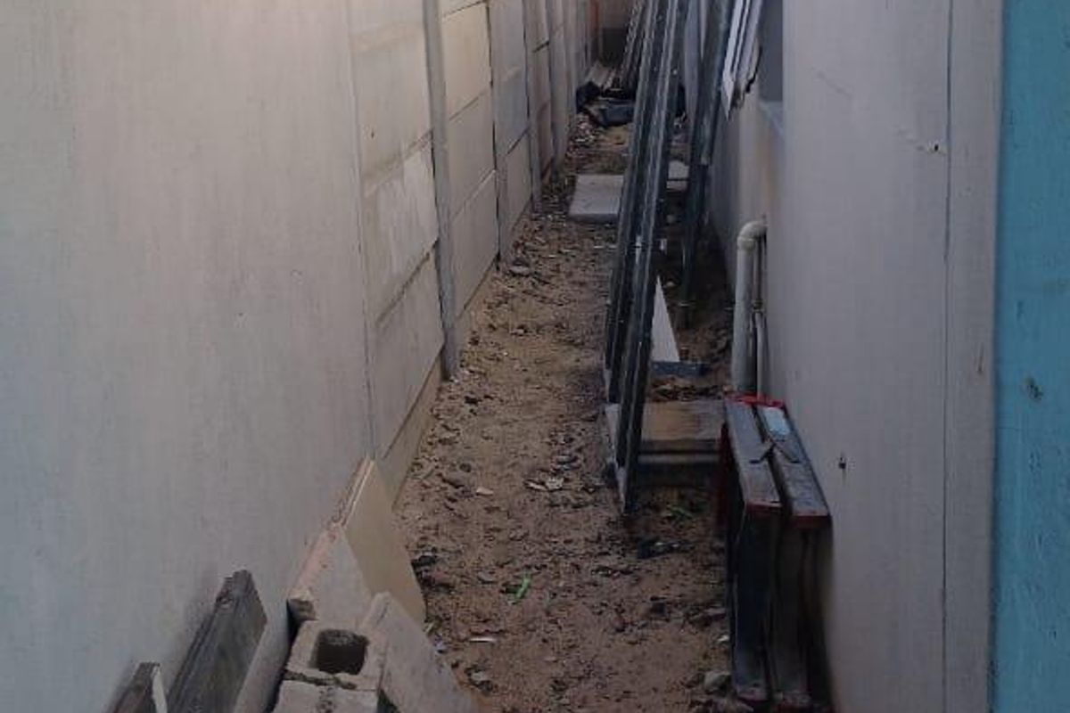 A narrow side passage between the house and a precast concrete boundary wall, currently unpaved and containing construction debris.