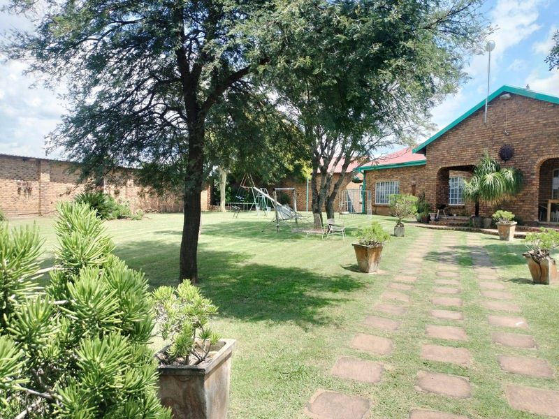 A spacious rear garden featuring a well-maintained lawn, mature trees, a paved walkway, and a brick boundary wall.
