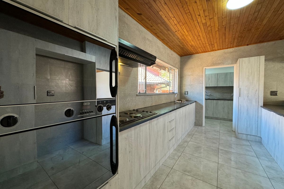 A modern kitchen featuring light-toned cabinetry, built-in oven and hob, tiled flooring, and a timber-clad ceiling, with an adjacent scullery area visible.