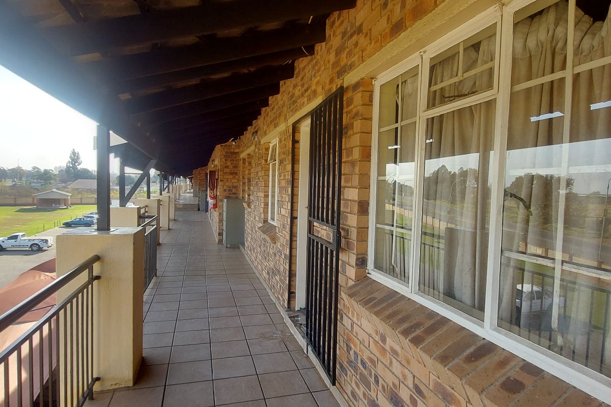 A covered balcony featuring tiled flooring, exposed timber rafters, and a brick facade, overlooking the complex grounds.