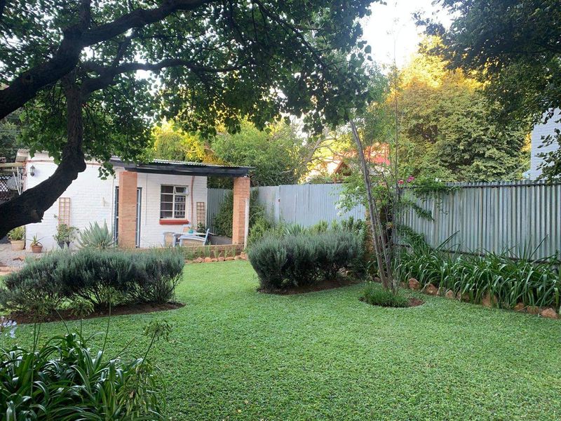 A well-maintained garden featuring a lush lawn, mature trees, and a covered patio area with brick pillars and corrugated metal fencing.