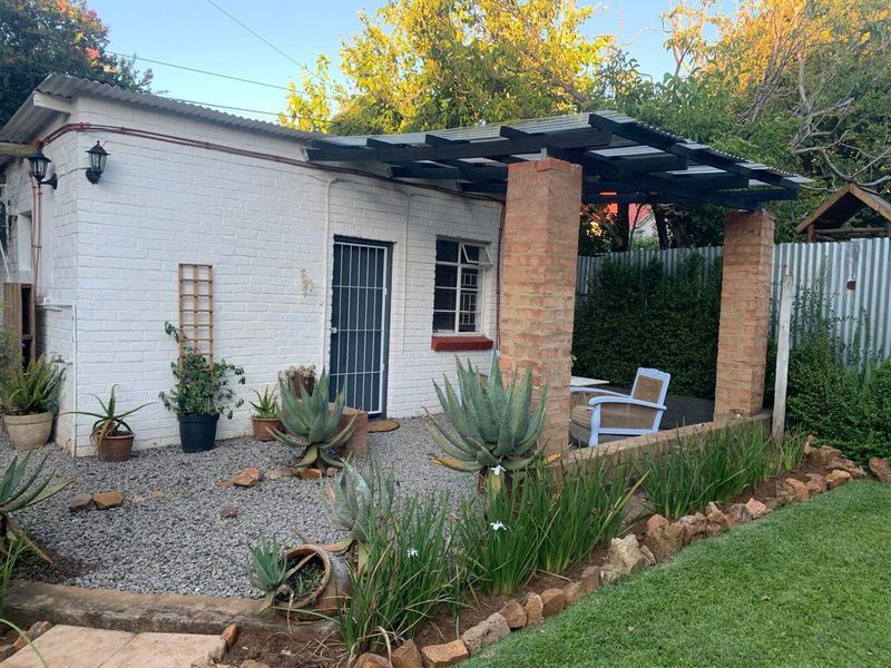A charming rear exterior view featuring a painted brick structure with a covered patio area, pergola, and landscaped garden surroundings.