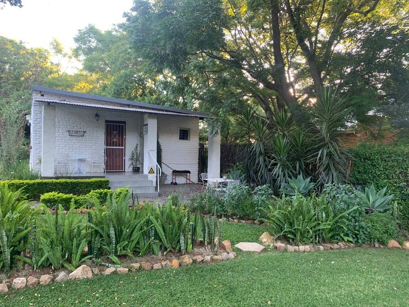 A charming cottage-style exterior featuring a painted brick facade, a covered patio entrance, and a well-maintained landscaped garden with mature trees.