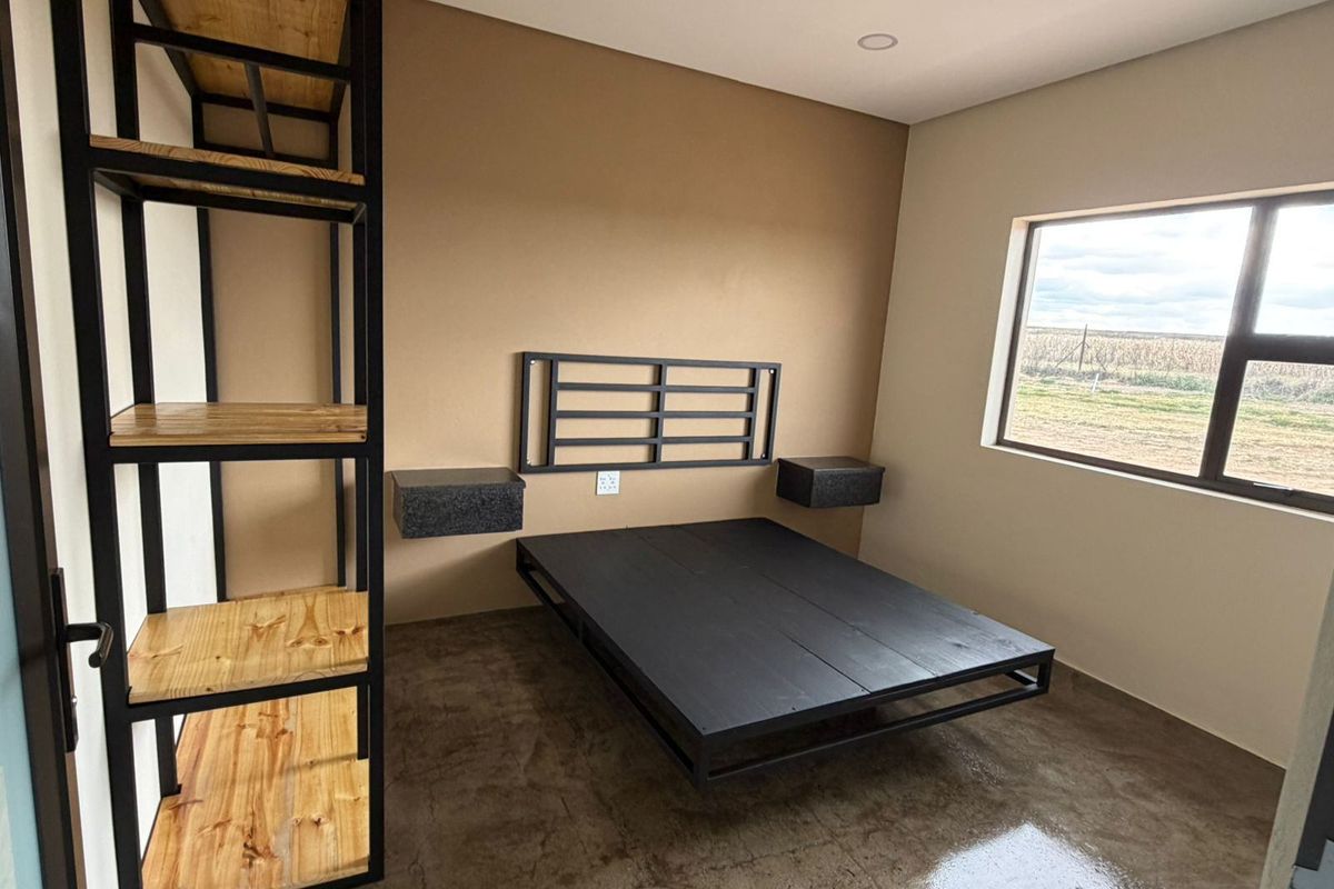A modern bedroom featuring polished concrete flooring, built-in metal shelving, floating bedside tables, and a large window with rural views.