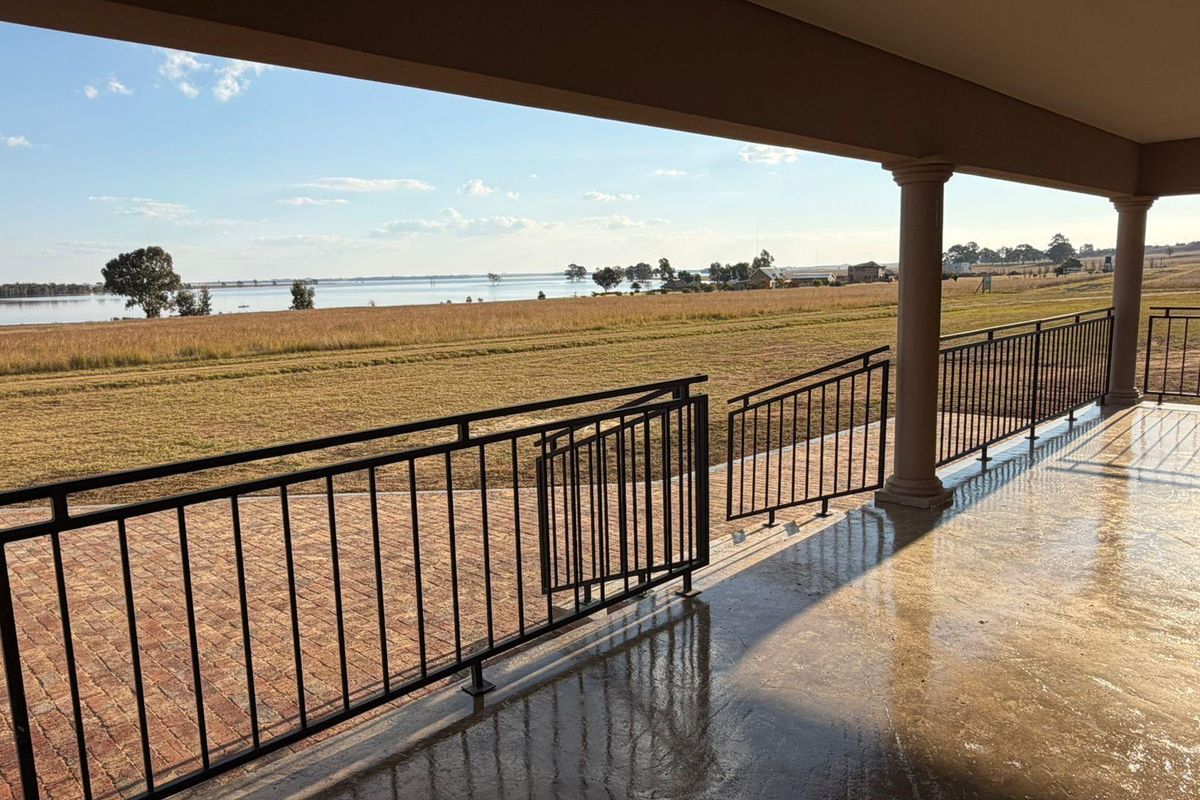 A spacious covered patio featuring a metal balustrade and brick paving, offering expansive views over the surrounding landscape and water.
