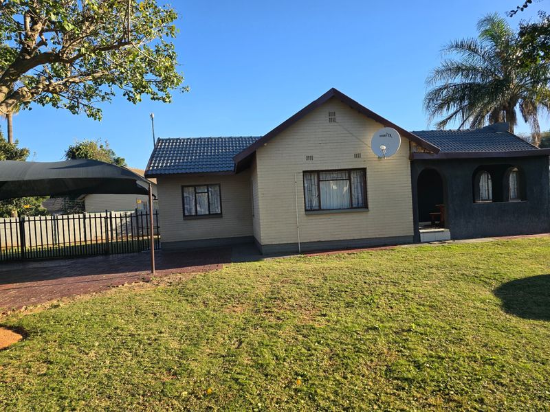 Front view of a single-story brick house featuring a tiled roof, paved driveway, and a shade cloth carport set within a grassed yard.