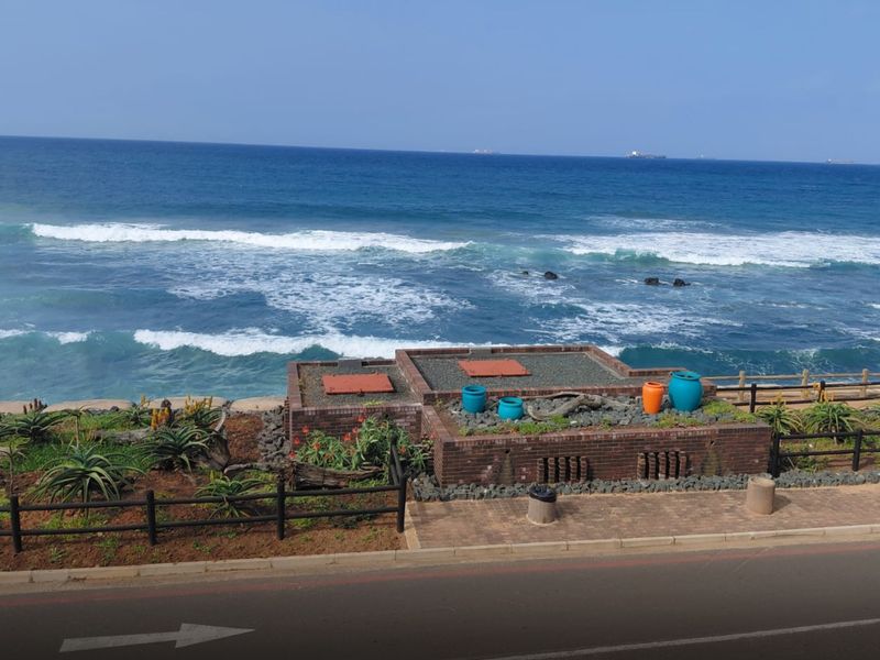 A scenic elevated view of the ocean coastline featuring a brick retaining wall, landscaped garden area, and a paved pedestrian walkway.