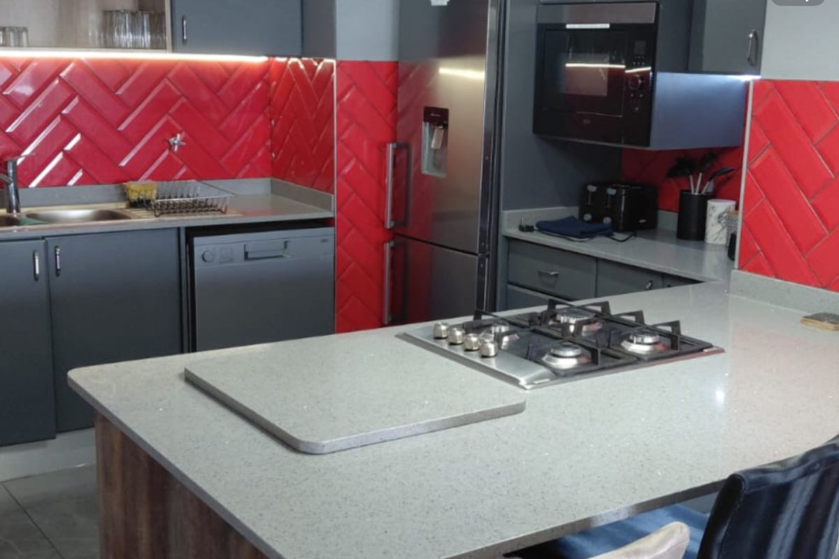 Modern kitchen featuring a central island with a gas hob, stone countertops, striking red herringbone tile backsplash, and integrated appliances.