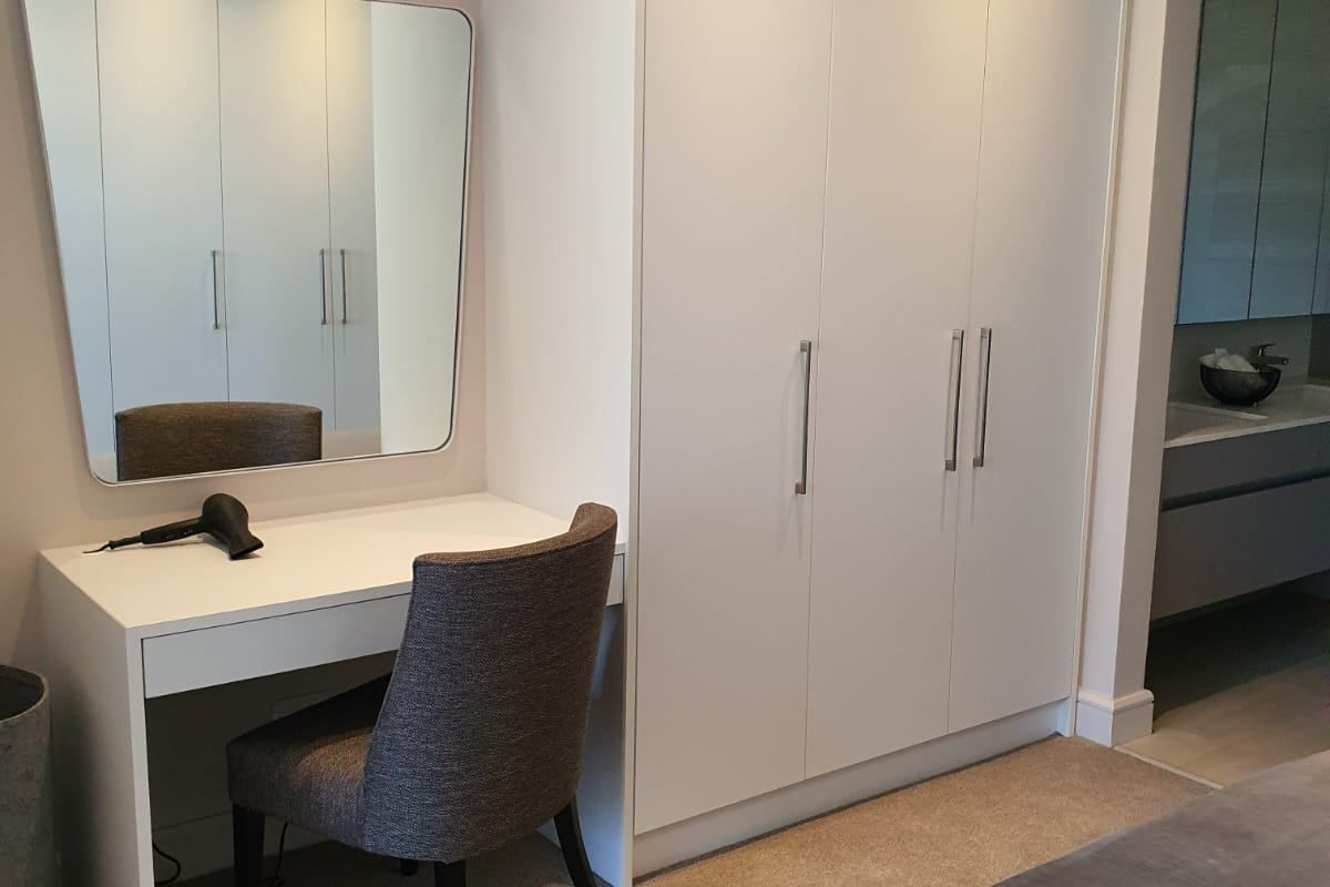 A well-maintained bedroom featuring a built-in wardrobe, integrated vanity desk, and recessed ceiling lighting, with an adjacent view into the bathroom area.