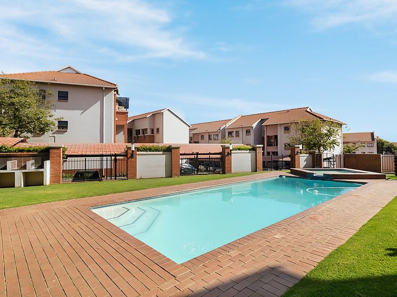 Communal swimming pool area featuring brick paving, manicured lawn, and built-in braai facilities within a secure apartment complex.