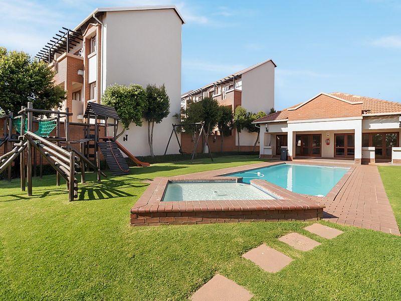 Communal outdoor area featuring a swimming pool, wading pool, brick-paved surround, and playground equipment set within a landscaped garden.