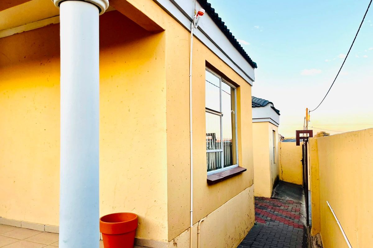 Side exterior view of the property featuring a covered patio area, paved walkway, and boundary wall with security gate.