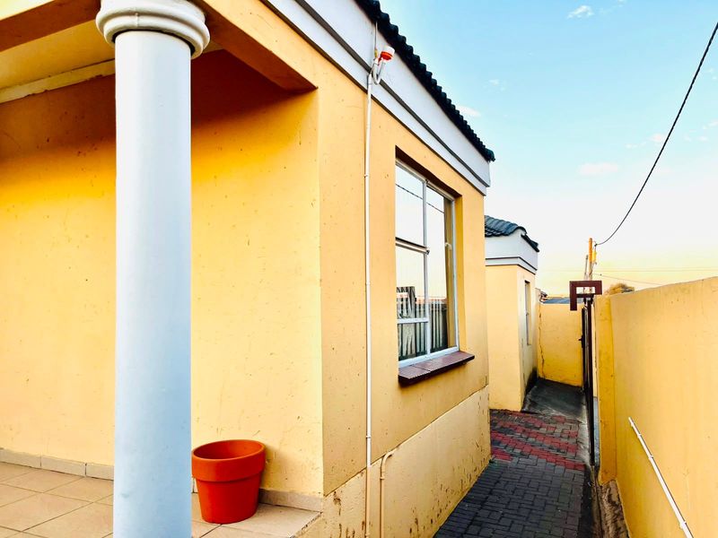 Side exterior view of the property featuring a covered patio area, paved walkway, and boundary wall with security gate.