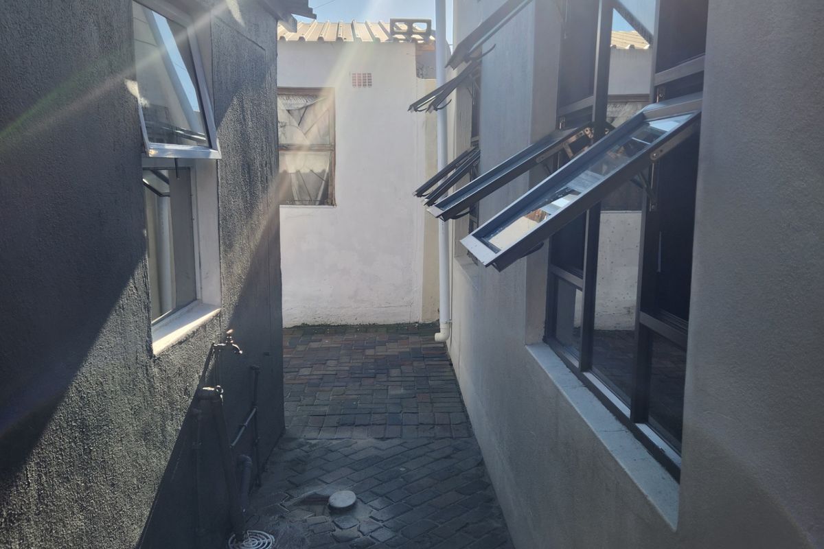 A narrow paved side passage between two building structures featuring external plumbing and multiple top-hung windows.