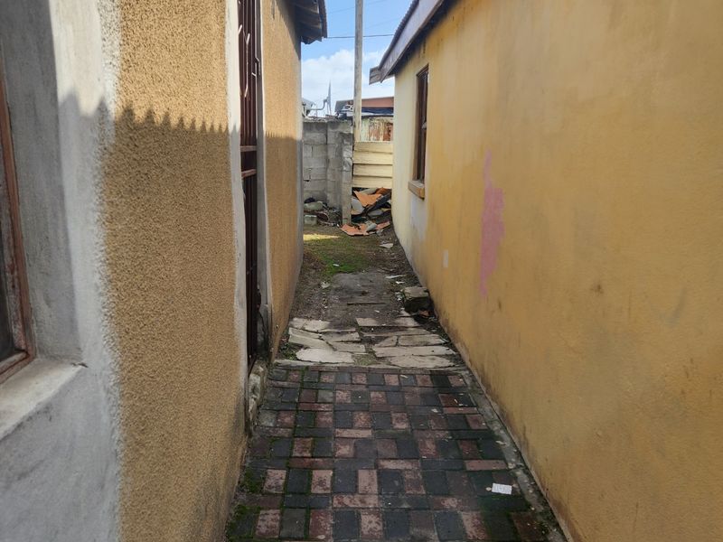A narrow side passage between two structures featuring paved flooring, security gate access, and boundary walls requiring maintenance.