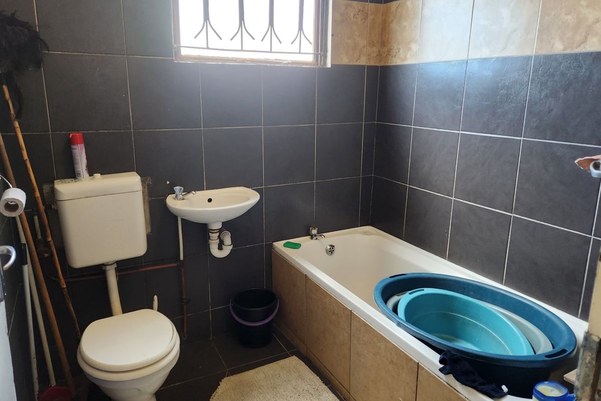 A compact bathroom featuring dark wall tiles, a standard bathtub, a wall-mounted basin, and a toilet, requiring some cosmetic updates to the finishes.