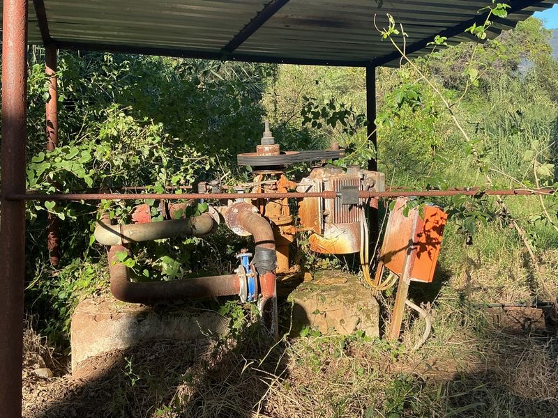 Outdoor water pump station featuring an electric motor and piping, protected by a simple metal roof structure on a concrete base.