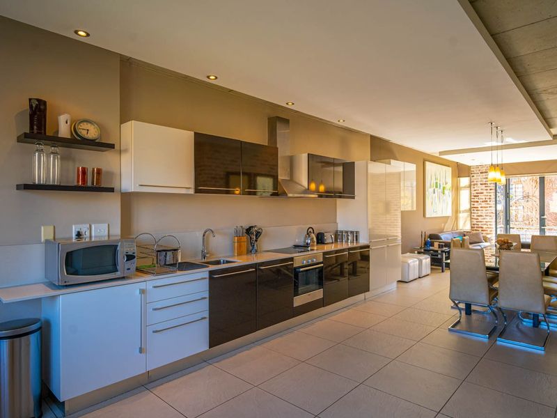 Modern open-plan kitchen and dining area featuring sleek cabinetry, integrated appliances, tiled floors, and an industrial-style exposed concrete ceiling and brick wall.