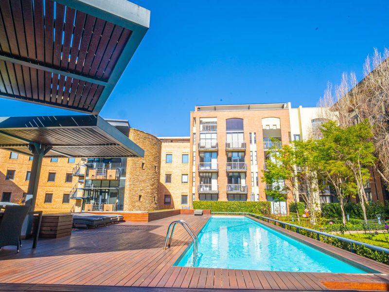A well-maintained communal pool area featuring timber decking, a modern metal pergola, and landscaped garden surrounds within an apartment complex.