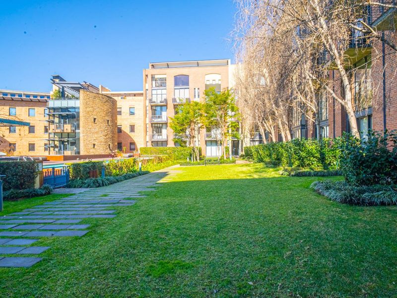 A well-maintained communal garden area featuring a stone pathway, manicured lawn, and views of the apartment complex brick facade and swimming pool.
