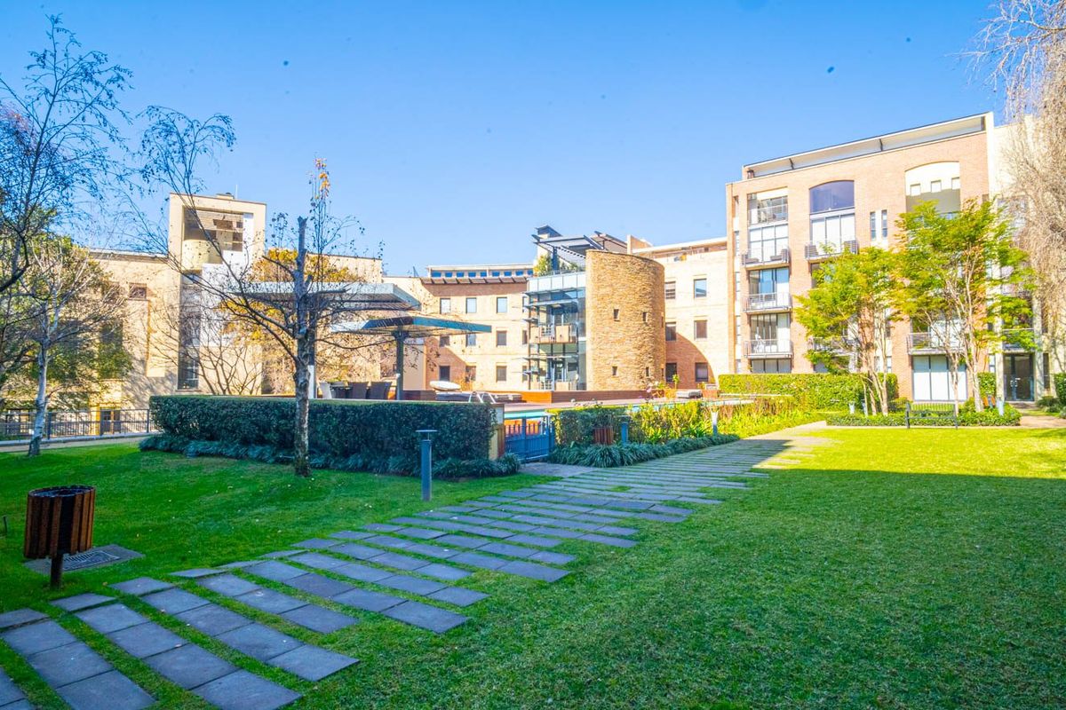 A well-maintained communal garden area featuring a paved walkway, landscaped lawns, and a swimming pool set against the apartment building facade.