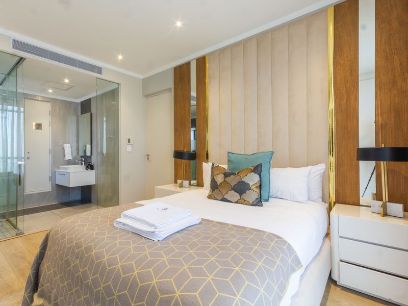 Modern bedroom featuring a custom upholstered headboard with integrated mirrors, recessed lighting, and a sleek glass-enclosed ensuite bathroom with a wall-mounted vanity.