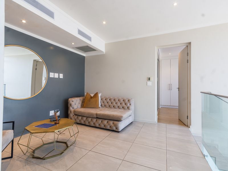 A modern living area featuring tiled floors, ducted air conditioning, recessed lighting, and a glass balustrade, leading to a hallway with built-in storage.