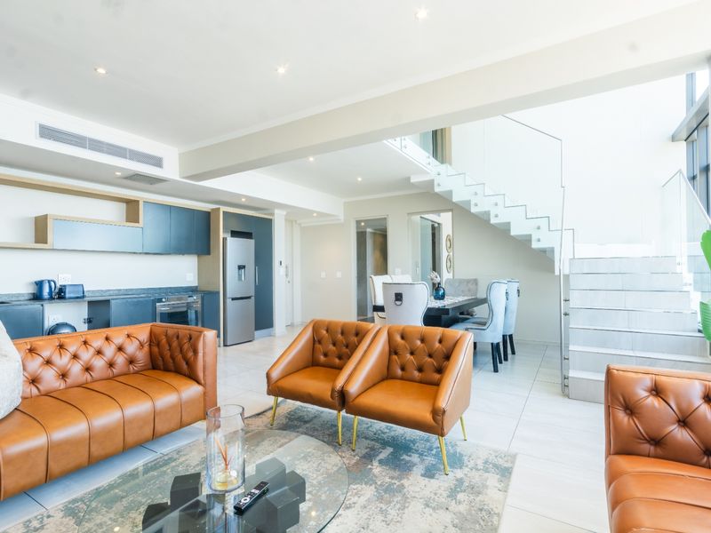 Modern open-plan living area featuring a sleek kitchen, tiled floors, recessed lighting, and a contemporary staircase with a glass balustrade.