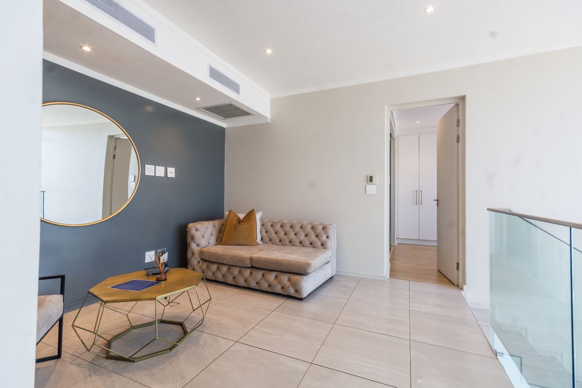 A modern living area featuring tiled floors, ducted air conditioning, recessed lighting, and a glass balustrade, leading to a hallway with built-in storage.