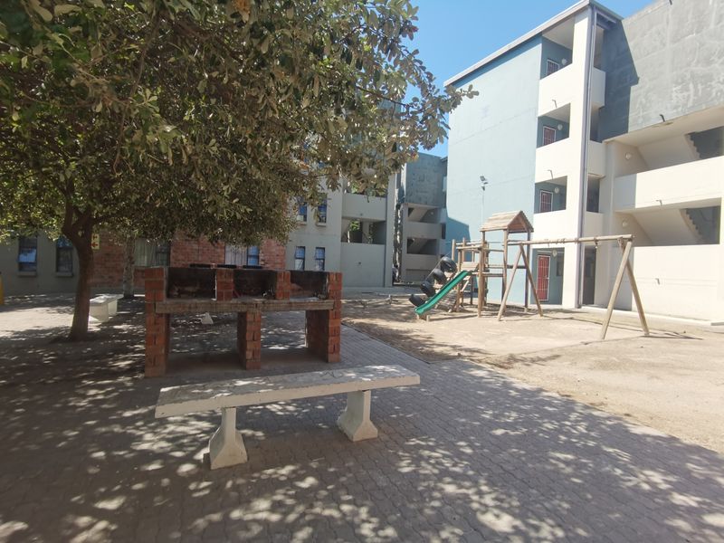 Communal outdoor area featuring a brick braai, concrete bench, and children's playground equipment set within a paved courtyard between apartment blocks.